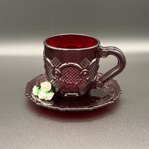 May include: A deep red glass teacup and saucer set featuring a raised diamond pattern and circular motifs. The cup has a curved handle with textured detailing, and the saucer has a scalloped edge. Small white artificial flowers rest on the saucer.