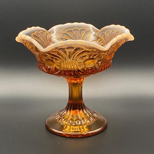 May include: An amber-colored, pedestal-style glass bowl. The bowl has a scalloped rim and is decorated with a floral pattern. The base is round and the stem is fluted. The glass has a glossy finish.