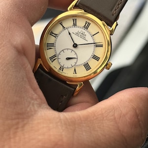Peut inclure: Une montre-bracelet Club La Mer dorée avec un cadran blanc, des chiffres romains noirs et un petit cadran secondaire. La montre a un bracelet en cuir marron et est tenue par une main. Le cadran de la montre indique "Club La Mer Urban Traditional For Exciting Professionals."