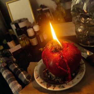 May include: A lit, red apple-shaped candle on a small white plate. The candle is dripping with wax. A small, plaid doll is in the foreground. Other items in the background include a skull-shaped bottle, small bottles of essential oils, and a mirror.