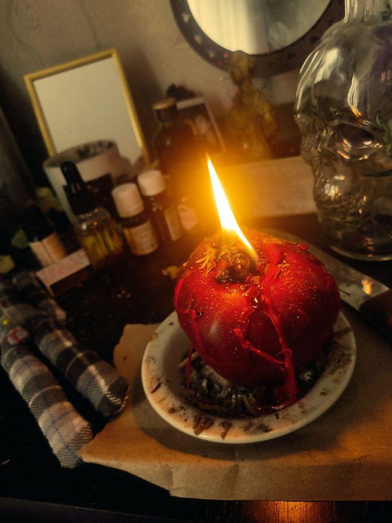 May include: A lit, red apple-shaped candle on a small white plate. The candle is dripping with wax. A small, plaid doll is in the foreground. Other items in the background include a skull-shaped bottle, small bottles of essential oils, and a mirror.