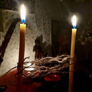 May include: Two lit, slender, yellow candles with bright flames. The candles are tied together with a length of twine. The background is dark, with a blurred image of a figure and a red object.