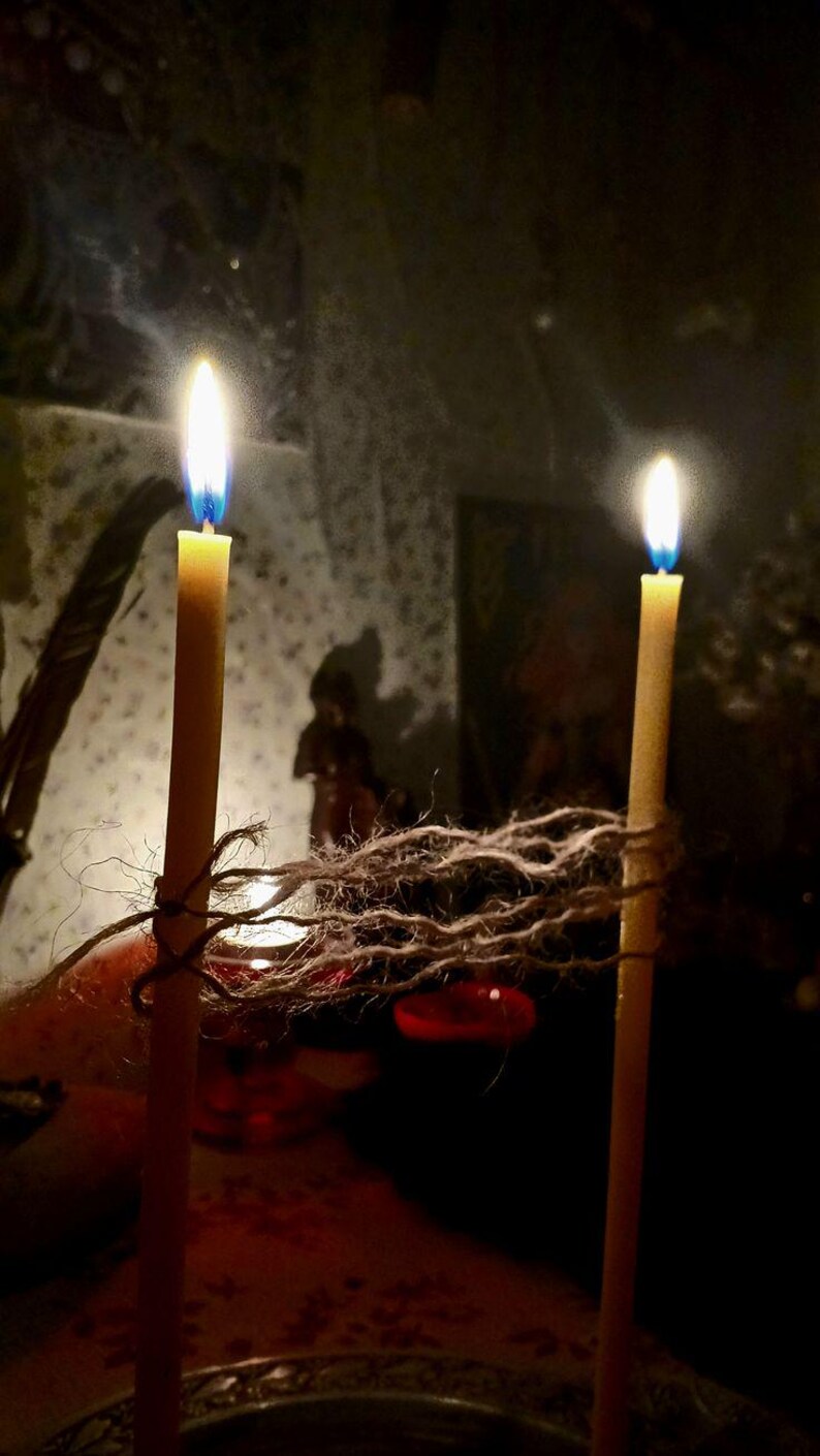 May include: Two lit, slender, yellow candles with bright flames. The candles are tied together with a length of twine. The background is dark, with a blurred image of a figure and a red object.