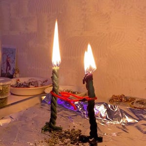 May include: Two lit, twisted candles with green and black wax, connected by red string. The candles sit on a foil-covered surface, with a small dish and other items in the background. The flames are bright and the scene has a mystical feel.