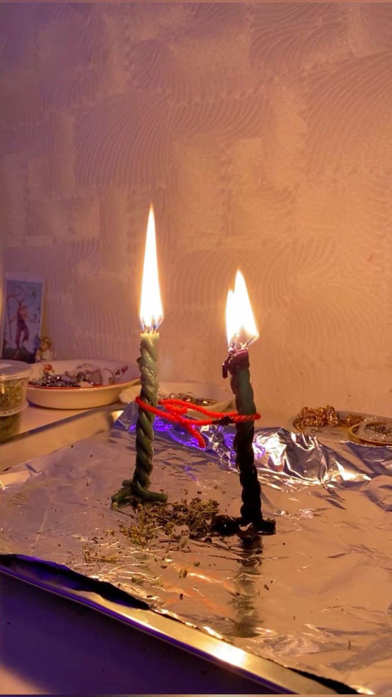 May include: Two lit, twisted candles with green and black wax, connected by red string. The candles sit on a foil-covered surface, with a small dish and other items in the background. The flames are bright and the scene has a mystical feel.