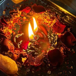 May include: A lit black candle with two flames, surrounded by red rose petals, herbs, and crystals on a silver tray. Two heart-shaped crystals are visible. The scene is illuminated with warm, inviting light.