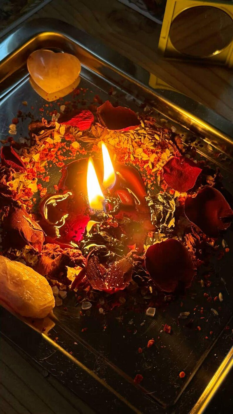May include: A lit black candle with two flames, surrounded by red rose petals, herbs, and crystals on a silver tray. Two heart-shaped crystals are visible. The scene is illuminated with warm, inviting light.