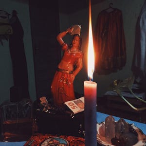 May include: A lit candle with a long flame stands in front of a red-orange statue of a woman. A deck of cards, a small box, and a plate of crystals are on a blue surface. A 50-dollar bill is visible.
