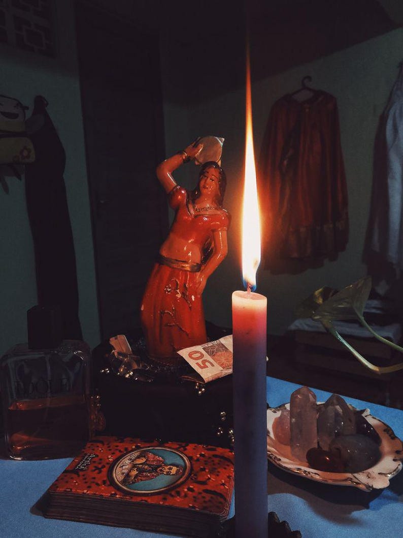 May include: A lit candle with a long flame stands in front of a red-orange statue of a woman. A deck of cards, a small box, and a plate of crystals are on a blue surface. A 50-dollar bill is visible.