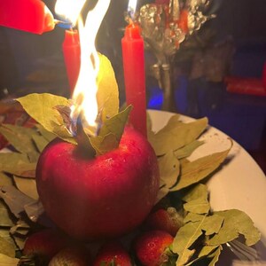 May include: A ritualistic arrangement featuring a red apple with a burning flame, surrounded by bay leaves and strawberries. Several red candles are lit, with one being lit by another. The scene is set on a white plate.