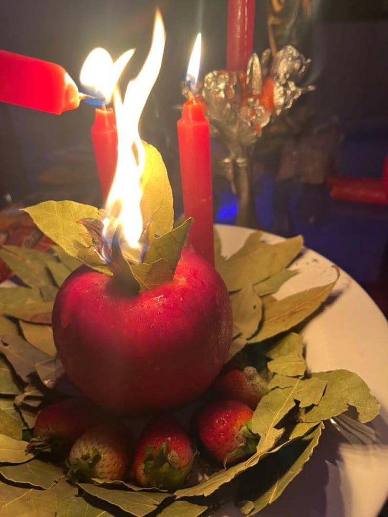 May include: A ritualistic arrangement featuring a red apple with a burning flame, surrounded by bay leaves and strawberries. Several red candles are lit, with one being lit by another. The scene is set on a white plate.