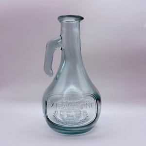 May include: A clear glass olive oil bottle with a handle. The bottle has a rounded body and a long neck. The words "OLIO EXTRAVERGINE D'OLIVA" are embossed on the front of the bottle. The bottle is empty.