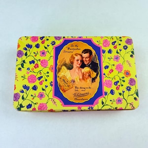 Könnte beinhalten: Vintage Whitman's Chocoladen-Blechdose mit gelbem Hintergrund und Blumenmuster. Die Dose zeigt ein zentrales Bild eines Paares und den Text "For the Particular Someone". Rechteckige Dose.