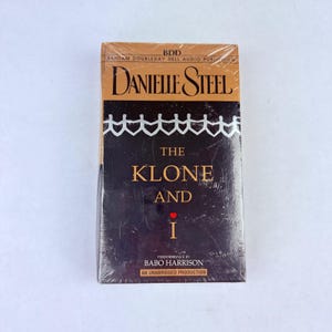 Puede incluir: Un audiolibro sellado de Danielle Steel, titulado "The Klone and I". La portada presenta un diseño dorado y negro con el nombre del autor en la parte superior. El título está en letras blancas grandes, con un símbolo de corazón rojo.