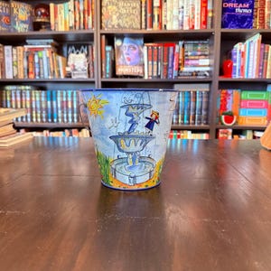 May include: A hand-painted, blue and white metal bucket with a whimsical fountain design. The bucket features a sunny sky, a fountain with a figure, and a handle. The bucket is sitting on a dark wooden table in front of a bookshelf.