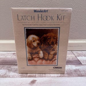 Op de afbeelding: Een WonderArt-haakpakket met een ontwerp van twee golden retriever-puppy's. De kit bevat de tekst "Latch Hook Kit" en "Tapissserie au Crochet". De doos is beige en bevat de tekst "Tool Not Included". Geschatte afmetingen: 50,8 cm x 68,6 cm.