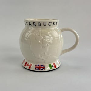 May include: Cream-colored Starbucks mug with a globe design. The mug features the word "STARBUCKS" in black lettering. The base of the mug is decorated with flags. The mug has a rounded shape and a curved handle.