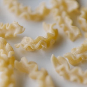 May include: Close-up of uncooked mafaldine pasta. The pasta is a light yellow color and has a ruffled, ribbon-like shape. Several pieces are scattered across a white surface, with some in focus and others blurred.