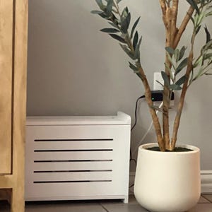 May include: A white storage cabinet with horizontal slats is positioned next to a large white planter holding a faux olive tree. The cabinet is rectangular and sits on the floor. The tree has brown branches and green leaves.