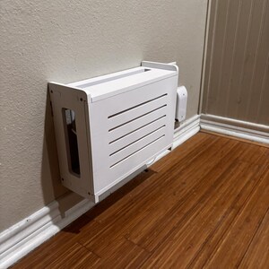 Puede incluir: Una cubierta blanca para caja de cables montada en la pared con listones horizontales en la parte delantera. La cubierta es rectangular y tiene una abertura superior. Est&aacute; montada en una pared beige con suelo de madera.