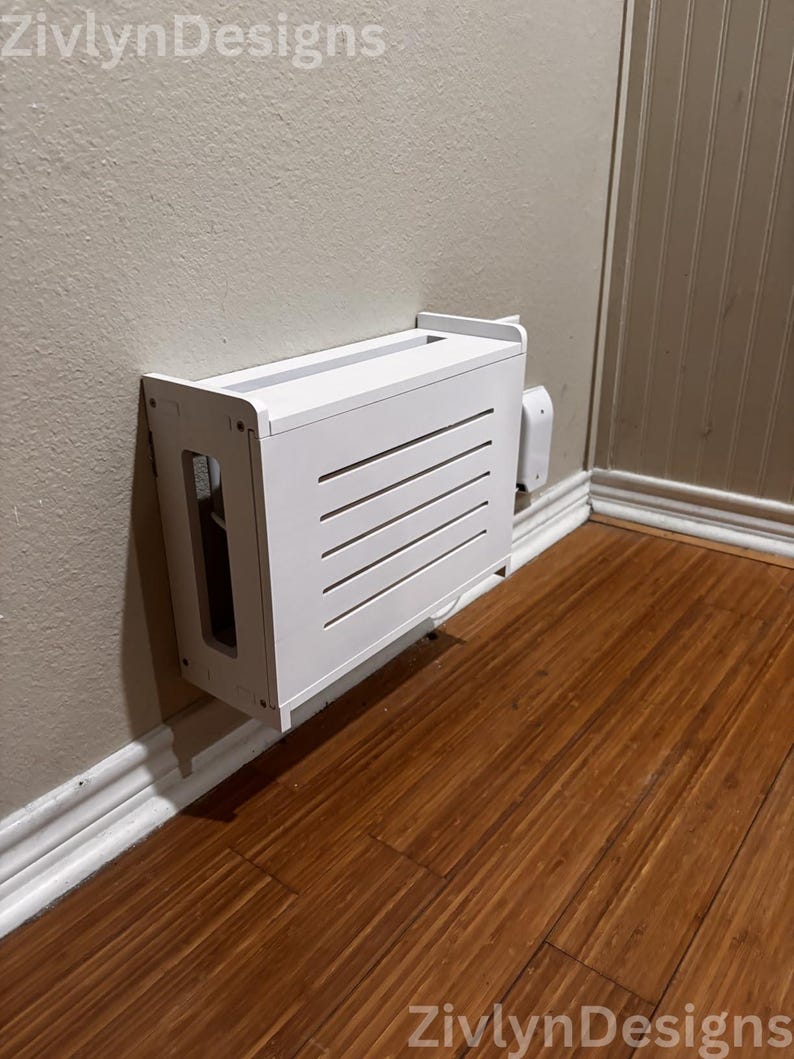 Puede incluir: Una cubierta blanca para caja de cables montada en la pared con listones horizontales en la parte delantera. La cubierta es rectangular y tiene una abertura superior. Est&aacute; montada en una pared beige con suelo de madera.