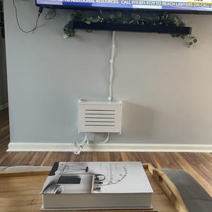 Puede incluir: Una escena de sala de estar con un televisor, una caja de cable blanca y una mesa de centro. Un libro y un mando a distancia est&aacute;n sobre la mesa. El televisor muestra el texto "WRAL NEWS 4:05". La caja de cable est&aacute; montada en la pared.