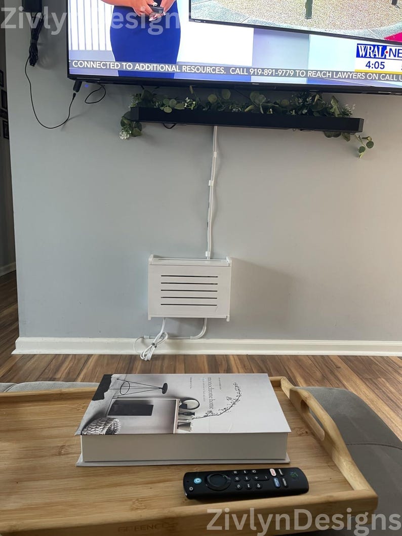 Puede incluir: Una escena de sala de estar con un televisor, una caja de cable blanca y una mesa de centro. Un libro y un mando a distancia est&aacute;n sobre la mesa. El televisor muestra el texto "WRAL NEWS 4:05". La caja de cable est&aacute; montada en la pared.