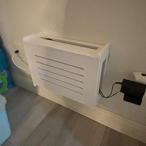 Puede incluir: Una caja blanca rectangular montada en la pared con listones horizontales en la parte delantera. La caja est&aacute; montada en una pared de color claro y tiene un adaptador de corriente enchufado a una toma de corriente. La marca "ZivlynDesigns" es visible.