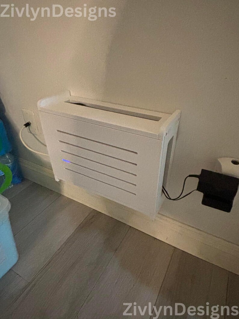 Puede incluir: Una caja blanca rectangular montada en la pared con listones horizontales en la parte delantera. La caja est&aacute; montada en una pared de color claro y tiene un adaptador de corriente enchufado a una toma de corriente. La marca "ZivlynDesigns" es visible.