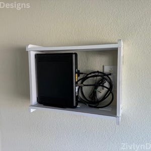 Puede incluir: Una unidad de estanter&iacute;a blanca, montada en la pared, alberga un dispositivo electr&oacute;nico negro con m&uacute;ltiples puertos y un haz de cables negros. La unidad est&aacute; montada en una pared de color claro.