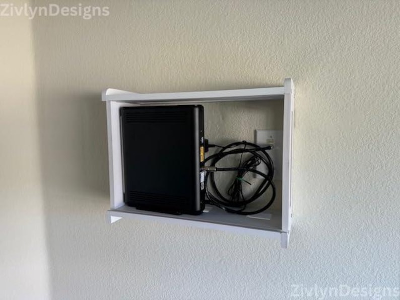 Puede incluir: Una unidad de estanter&iacute;a blanca, montada en la pared, alberga un dispositivo electr&oacute;nico negro con m&uacute;ltiples puertos y un haz de cables negros. La unidad est&aacute; montada en una pared de color claro.