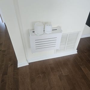 Puede incluir: Una cubierta blanca para ventilaci&oacute;n con un estante que sostiene dos dispositivos electr&oacute;nicos blancos. El estante tiene listones horizontales. La pared es blanca y el suelo es de madera marr&oacute;n oscuro. El texto "ZivlynDesigns" est&aacute; en las esquinas superior izquierda e inferior derecha.