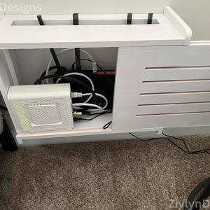 Puede incluir: Consola de medios de madera blanca con un compartimento incorporado para electr&oacute;nica. La consola cuenta con una balda superior, una puerta de lamas y un compartimento que alberga un m&oacute;dem, un router y varios cables. El dise&ntilde;o incluye el texto "ZivlynDesigns".