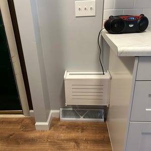 Puede incluir: Una cubierta de ventilaci&oacute;n rectangular blanca y una caja de ventilaci&oacute;n blanca a juego est&aacute;n montadas en una pared gris claro. Una radio negra y roja est&aacute; sobre una encimera blanca. El suelo es de laminado de madera oscura.