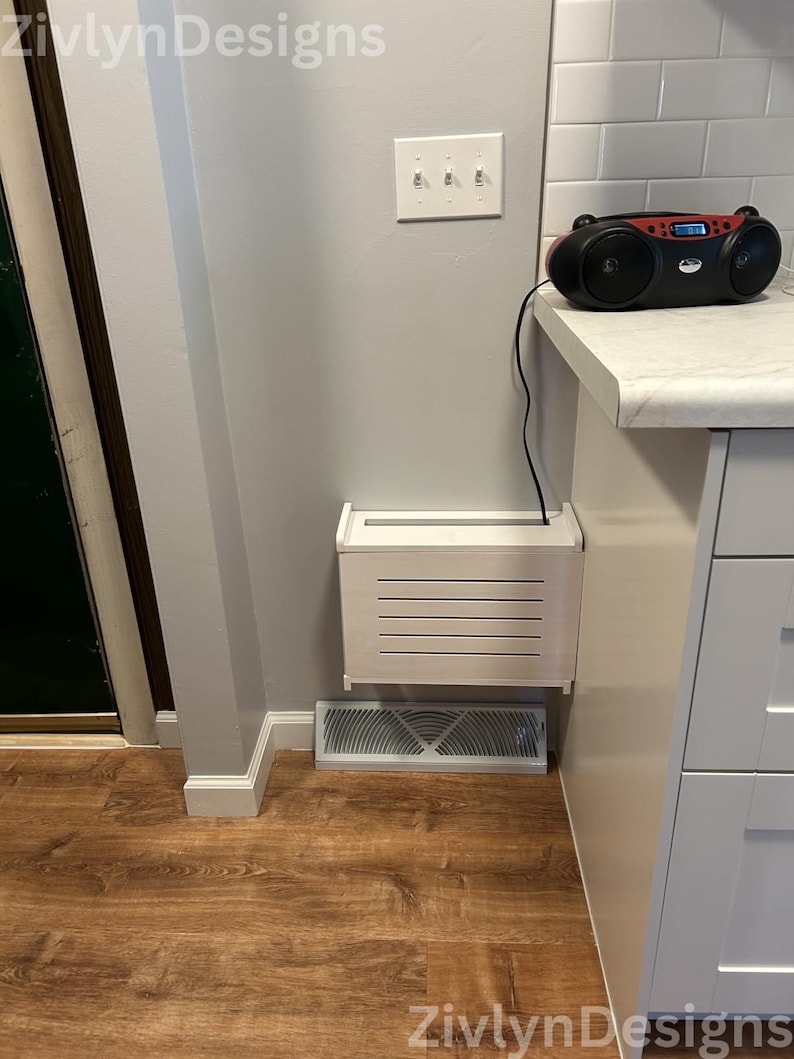 Puede incluir: Una cubierta de ventilaci&oacute;n rectangular blanca y una caja de ventilaci&oacute;n blanca a juego est&aacute;n montadas en una pared gris claro. Una radio negra y roja est&aacute; sobre una encimera blanca. El suelo es de laminado de madera oscura.