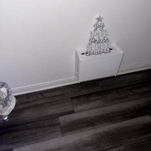 Puede incluir: Decoraci&oacute;n navide&ntilde;a blanca con forma de &aacute;rbol, con letras plateadas que dicen "Joy, Noel, Peace, Believe, Christmas". El &aacute;rbol est&aacute; montado sobre una caja blanca, ideal para la decoraci&oacute;n festiva del hogar.