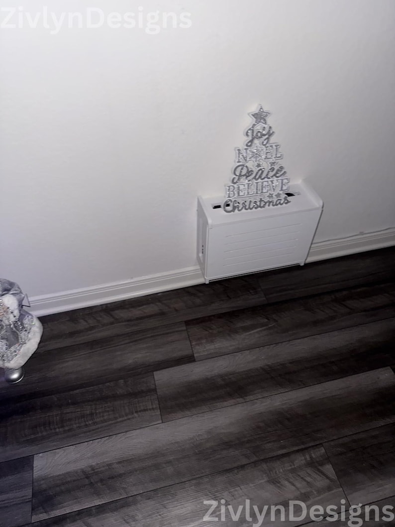 Puede incluir: Decoraci&oacute;n navide&ntilde;a blanca con forma de &aacute;rbol, con letras plateadas que dicen "Joy, Noel, Peace, Believe, Christmas". El &aacute;rbol est&aacute; montado sobre una caja blanca, ideal para la decoraci&oacute;n festiva del hogar.