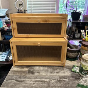 May include: Two-tiered bamboo bread box with clear, ribbed glass doors and a small wooden knob on each door. The bread boxes are stacked on a wooden table with a gray wood grain pattern.