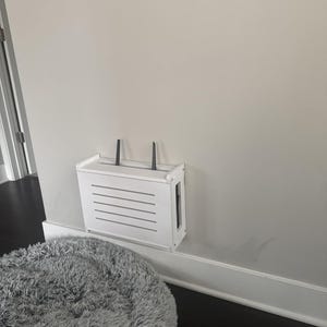 Puede incluir: Un recinto blanco para enrutador montado en la pared con una parte delantera con listones y dos antenas negras. El recinto est&aacute; montado en una pared gris claro sobre una cama para mascotas gris y esponjosa. La marca "ZivlynDesigns" est&aacute; en las esquinas superior izquierda e inferior derecha.