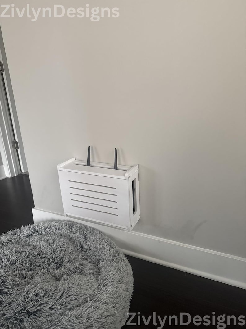 Puede incluir: Un recinto blanco para enrutador montado en la pared con una parte delantera con listones y dos antenas negras. El recinto est&aacute; montado en una pared gris claro sobre una cama para mascotas gris y esponjosa. La marca "ZivlynDesigns" est&aacute; en las esquinas superior izquierda e inferior derecha.