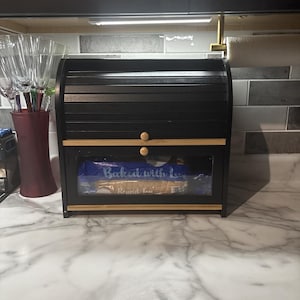 May include: Black wooden bread box with a roll-top door and a clear window. The bread box has a natural wood trim and two round wooden knobs. The text "Baked with Love" is visible through the window. The bread box is on a marble countertop.