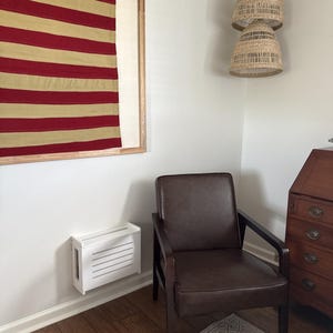 Puede incluir: Interior de una habitaci&oacute;n con una obra de arte enmarcada con rayas horizontales rojas y doradas. Un sill&oacute;n de cuero marr&oacute;n con marco de madera oscura se encuentra sobre una alfombra estampada. Tambi&eacute;n se ve una caja blanca montada en la pared y una l&aacute;mpara colgante tejida.