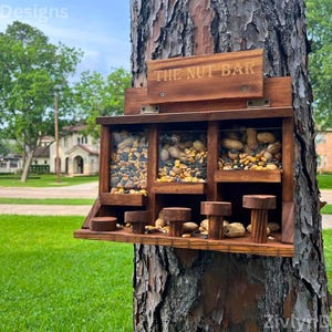 May include: A wooden bird feeder with three compartments, each filled with different types of birdseed and nuts. The feeder is attached to a tree trunk and has the words "THE NUT BAR" on top. The feeder is designed to attract birds.