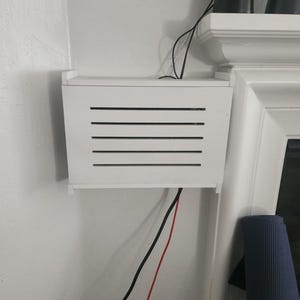 Puede incluir: Una caja rectangular blanca con listones negros horizontales montada en una pared blanca. Cables negros y rojos salen de la caja. El texto "ZivlynDesigns" es visible en las esquinas superior izquierda e inferior derecha.