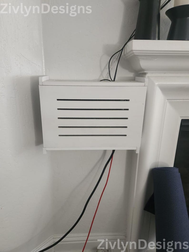 Puede incluir: Una caja rectangular blanca con listones negros horizontales montada en una pared blanca. Cables negros y rojos salen de la caja. El texto "ZivlynDesigns" es visible en las esquinas superior izquierda e inferior derecha.