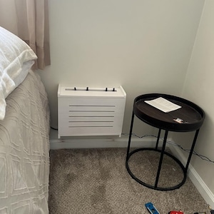 Puede incluir: Un mueble blanco rectangular con lamas horizontales, colocado junto a una cama y una peque&ntilde;a mesa auxiliar redonda. La mesa tiene una tapa marr&oacute;n oscuro y una estructura de metal negro. Herramientas en el suelo alfombrado.