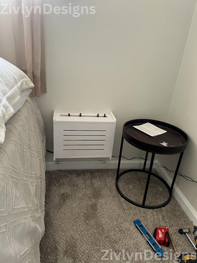 Puede incluir: Un mueble blanco rectangular con lamas horizontales, colocado junto a una cama y una peque&ntilde;a mesa auxiliar redonda. La mesa tiene una tapa marr&oacute;n oscuro y una estructura de metal negro. Herramientas en el suelo alfombrado.