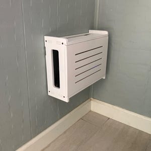 Puede incluir: Una caja blanca rectangular montada en la pared con una parte frontal ranurada y una gran abertura lateral. La caja est&aacute; montada en una pared gris en una esquina. El texto "ZivlynDesigns" est&aacute; en la parte superior izquierda e inferior derecha.