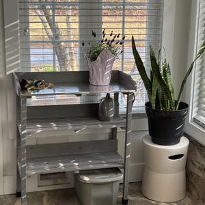 May include: A gray metal potting bench with two shelves and a top surface. A pink paper pot with flowers sits on the top shelf. A black potted plant sits on a white stool. The bench is near a window with blinds.