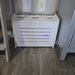 Puede incluir: Una caja de madera blanca con listones horizontales y una luz azul, dise&ntilde;ada para ocultar aparatos electr&oacute;nicos. La caja est&aacute; colocada sobre un suelo con aspecto de madera, con un enchufe de pared y un dispositivo blanco en el fondo.